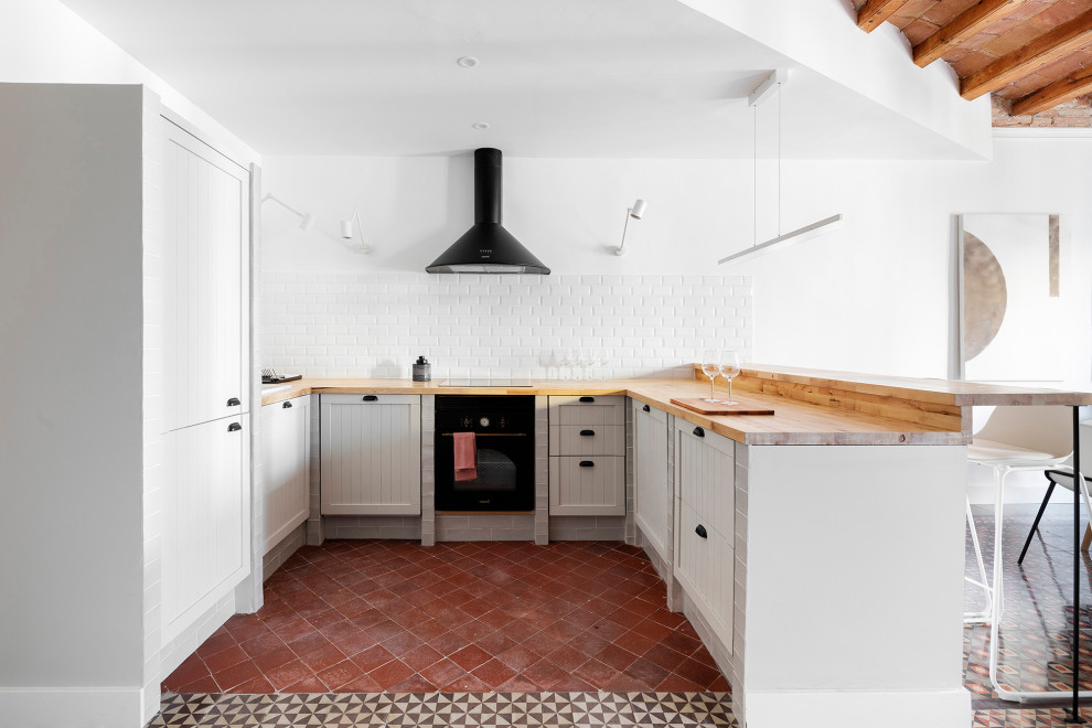 This is an example of a mediterranean u-shaped kitchen in Barcelona with a built-in sink, shaker cabinets, grey cabinets, wood worktops, white splashback, metro tiled splashback, black appliances, a breakfast bar, red floors and brown worktops.