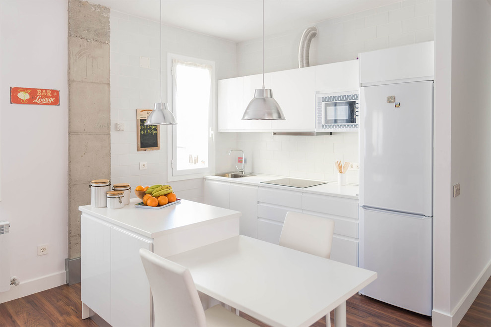 Exempel på ett litet industriellt linjärt kök och matrum, med en enkel diskho, släta luckor, vita skåp, vitt stänkskydd, vita vitvaror, mörkt trägolv och en köksö