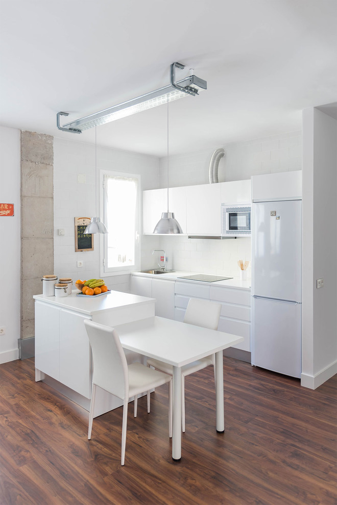 Bild på ett litet industriellt linjärt kök och matrum, med en enkel diskho, släta luckor, vita skåp, vitt stänkskydd, vita vitvaror, mörkt trägolv och en köksö