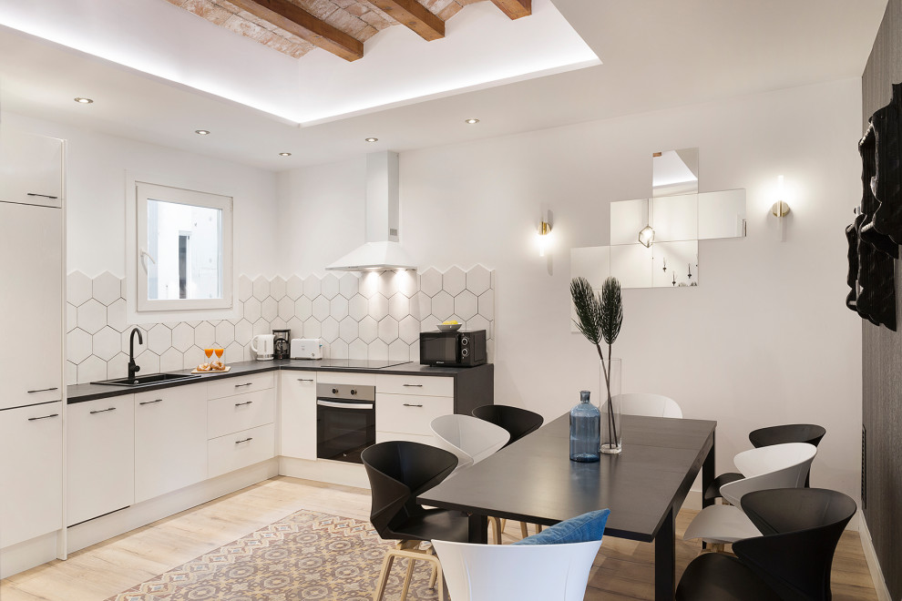 Mediterrane Wohnküche in L-Form mit Einbauwaschbecken, flächenbündigen Schrankfronten, weißen Schränken, Küchenrückwand in Weiß, hellem Holzboden, schwarzer Arbeitsplatte, freigelegten Dachbalken und eingelassener Decke in Barcelona