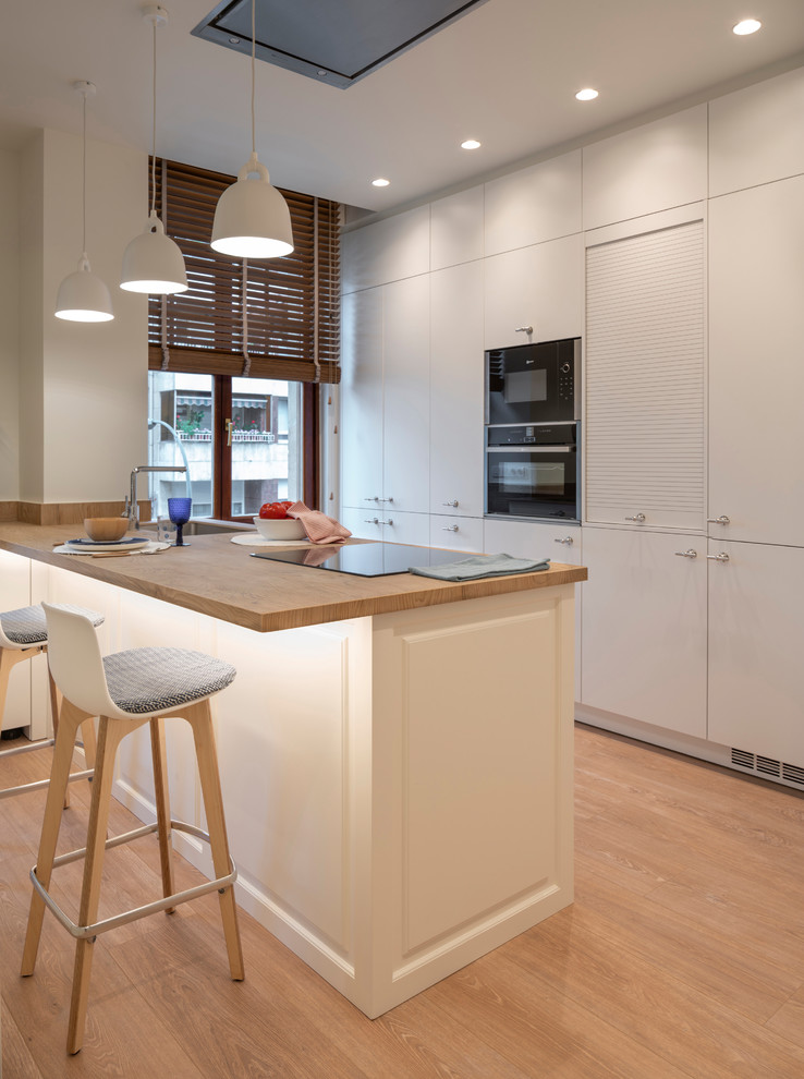 Modern inredning av ett stort brun brunt u-kök, med släta luckor, vita skåp, en halv köksö, en undermonterad diskho, integrerade vitvaror, brunt golv, träbänkskiva och ljust trägolv
