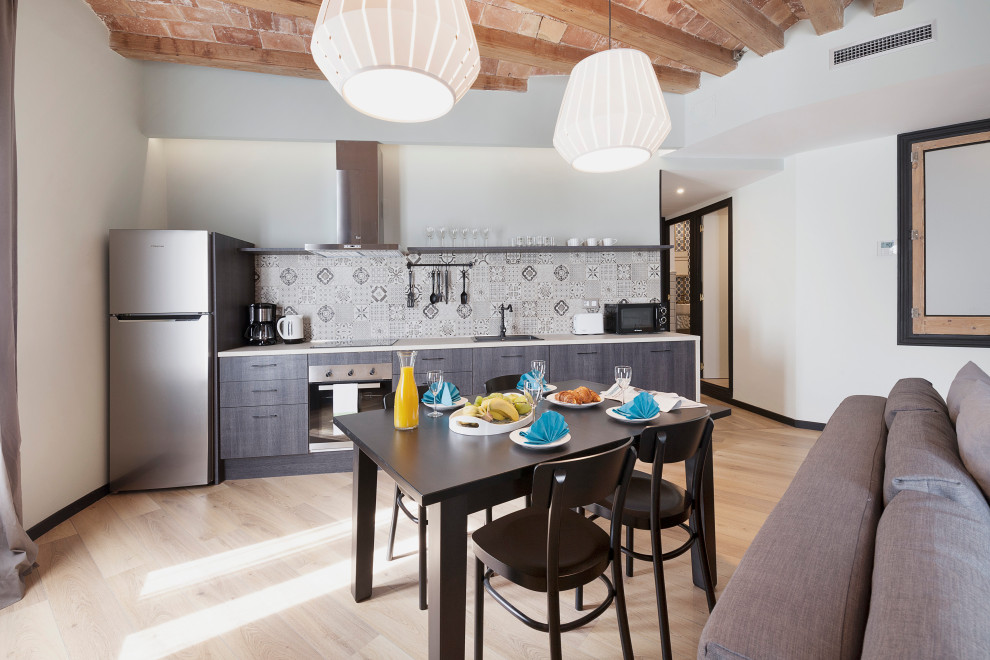 Example of an urban single-wall light wood floor and exposed beam open concept kitchen design in Barcelona with a drop-in sink, flat-panel cabinets, brown cabinets, multicolored backsplash, stainless steel appliances and white countertops