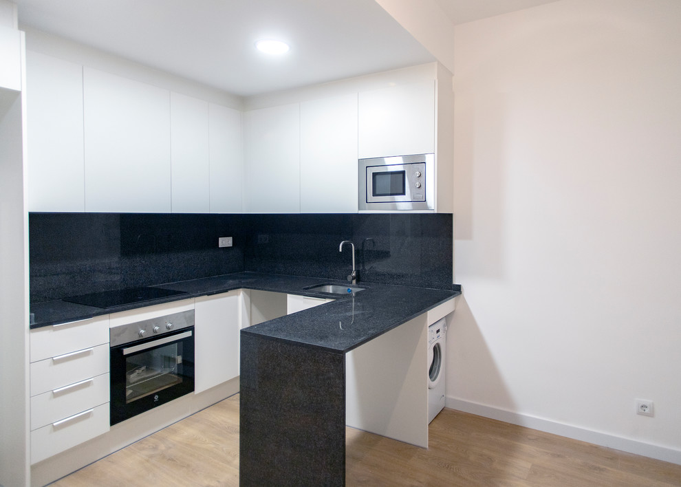 Photo of a medium sized contemporary u-shaped open plan kitchen in Barcelona with a single-bowl sink, flat-panel cabinets, white cabinets, black splashback, stainless steel appliances, medium hardwood flooring, no island, brown floors and black worktops.