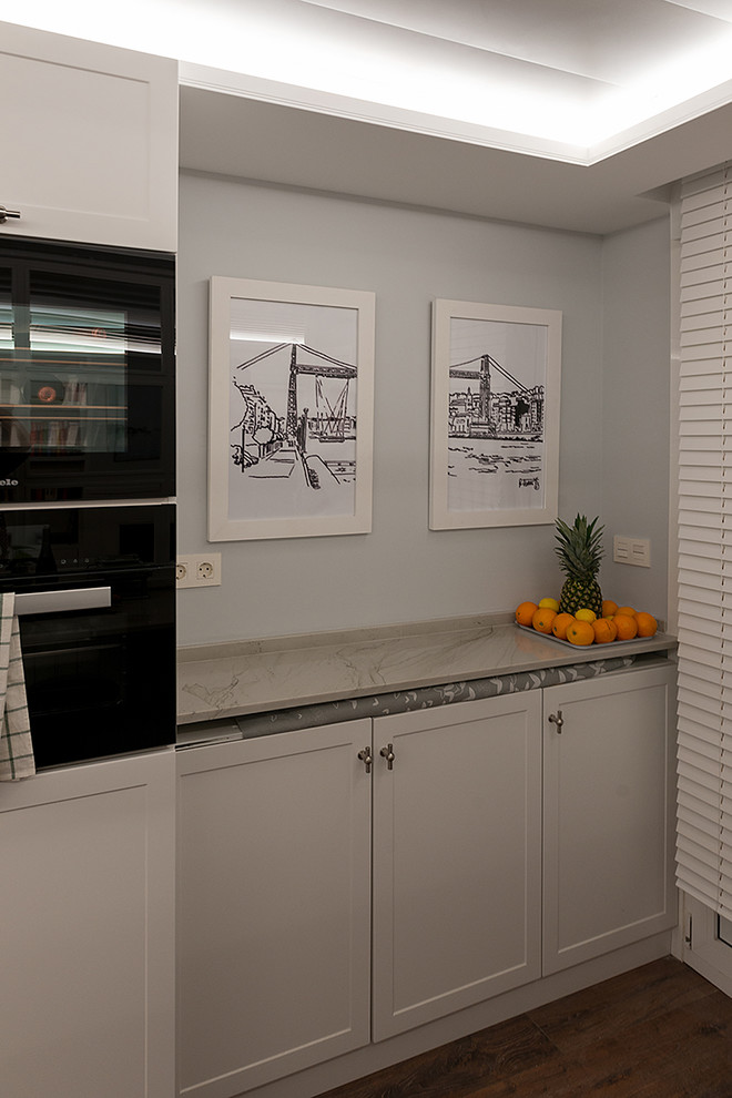 Offene Klassische Küche in L-Form mit Doppelwaschbecken, profilierten Schrankfronten, weißen Schränken, Granit-Arbeitsplatte, Kalk-Rückwand, schwarzen Elektrogeräten, braunem Holzboden und braunem Boden in Bilbao