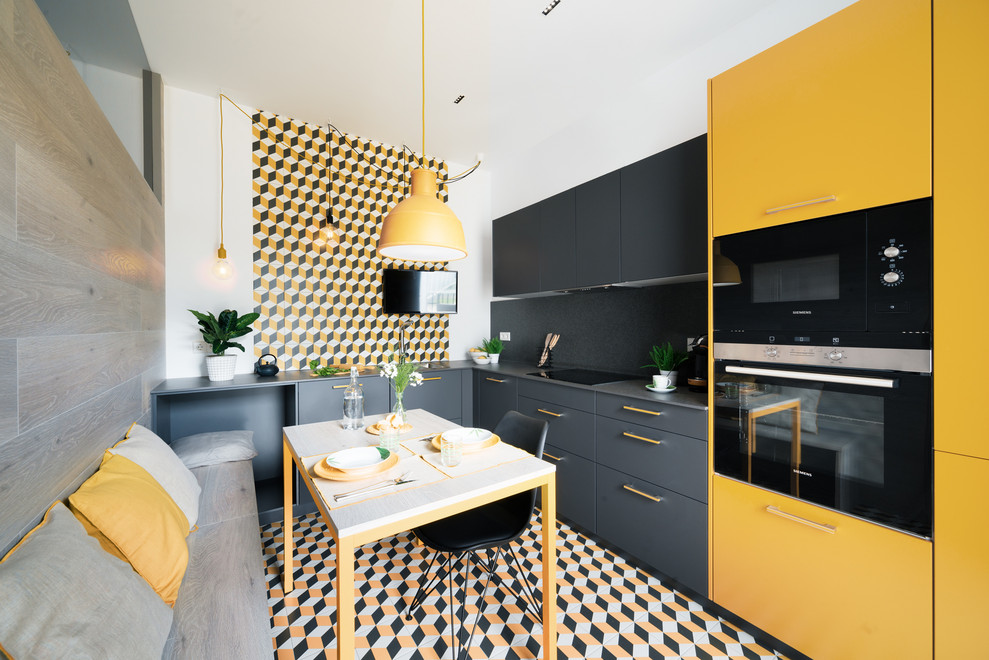 Cette image montre une cuisine américaine bicolore design en L avec un placard à porte plane, des portes de placard jaunes, une crédence multicolore, un électroménager noir, un sol multicolore et un plan de travail gris.