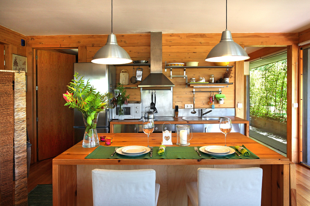 Aménagement d'une petite cuisine américaine linéaire exotique en inox avec un sol en bois brun, un évier posé, un placard à porte plane, un plan de travail en bois, une crédence métallisée, un électroménager en acier inoxydable, îlot et un sol marron.