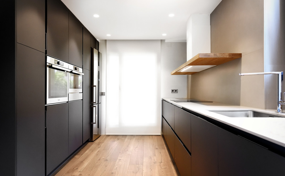 Ejemplo de cocina minimalista con fregadero bajoencimera, armarios con paneles lisos, puertas de armario negras, electrodomésticos de acero inoxidable, suelo de madera clara y encimeras blancas