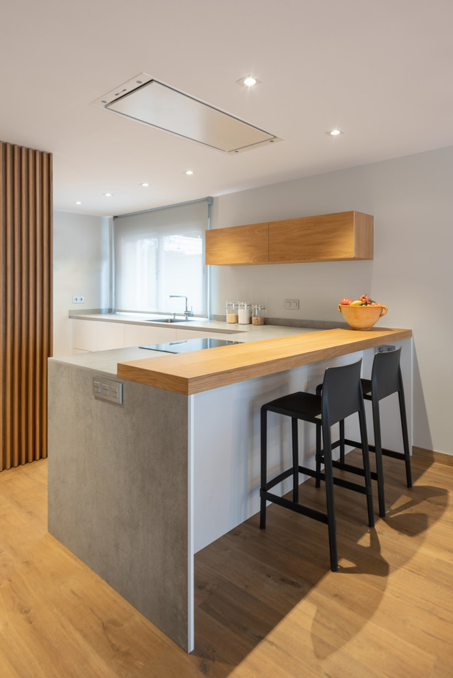 Moderne Küche in L-Form mit braunem Holzboden, Halbinsel, grauer Arbeitsplatte, flächenbündigen Schrankfronten, weißen Schränken und braunem Boden in Barcelona