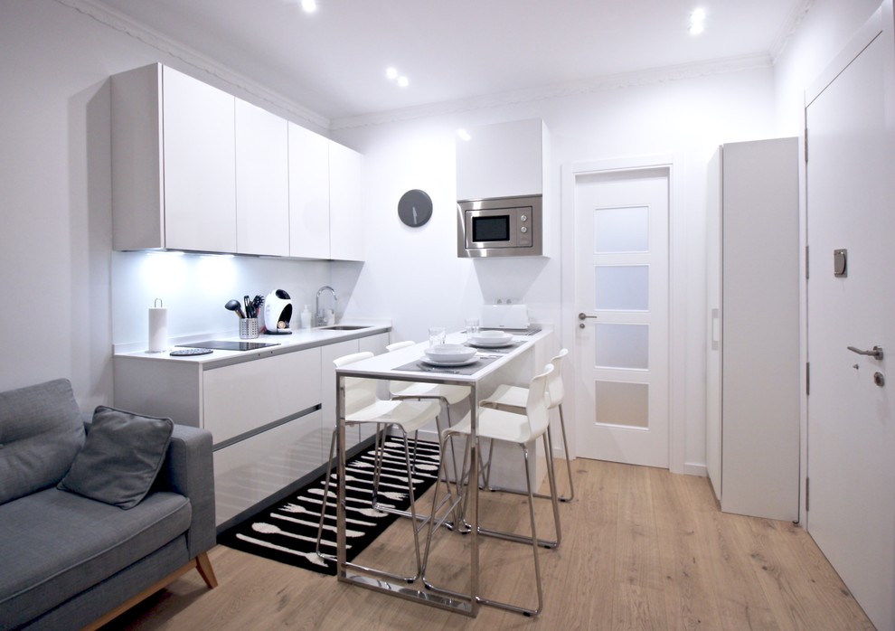 Example of a small danish light wood floor open concept kitchen design in Madrid with flat-panel cabinets, white cabinets, white backsplash, stainless steel appliances, a peninsula, white countertops and an undermount sink