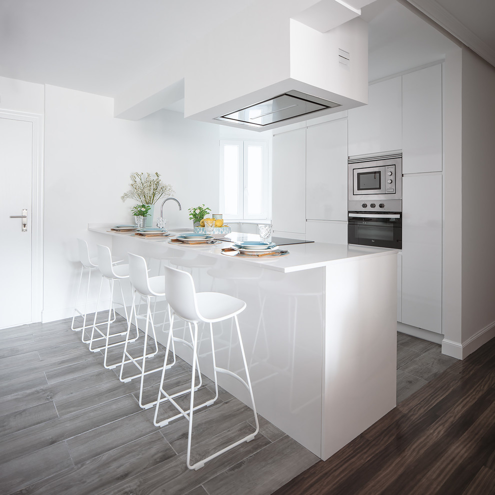 Zweizeilige Moderne Küche mit flächenbündigen Schrankfronten, weißen Schränken, Küchengeräten aus Edelstahl, Halbinsel, weißer Arbeitsplatte und grauem Boden in Bilbao