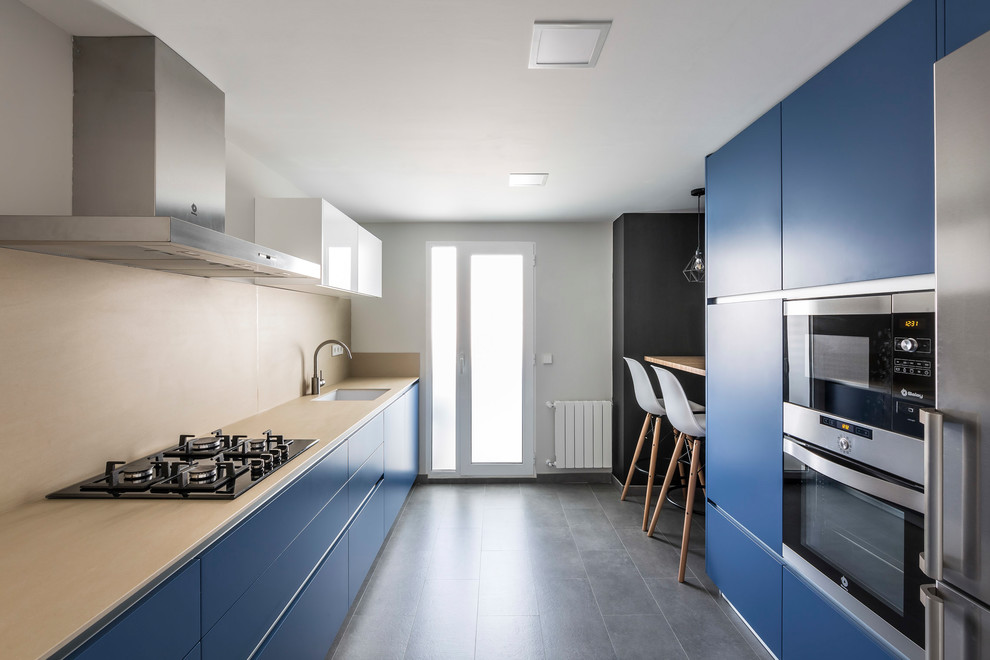 На фото: параллельная кухня в современном стиле с врезной мойкой, плоскими фасадами, синими фасадами, бежевым фартуком, техникой из нержавеющей стали и серым полом с