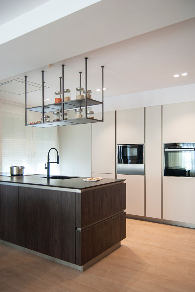 This is an example of a contemporary single-wall open plan kitchen in Bilbao with a submerged sink, flat-panel cabinets, beige cabinets, quartz worktops, stainless steel appliances, an island and black worktops.