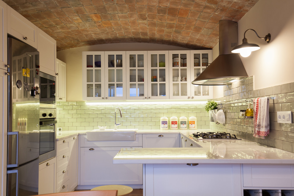 Inredning av ett klassiskt u-kök, med vita skåp, rostfria vitvaror, en halv köksö, en rustik diskho, luckor med glaspanel och stänkskydd i tunnelbanekakel