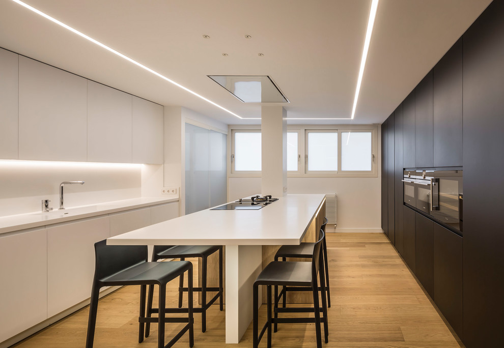This is an example of a large contemporary kitchen in Valencia with an integrated sink, flat-panel cabinets, white cabinets, engineered stone countertops, white splashback, black appliances, an island, white worktops and light hardwood flooring.