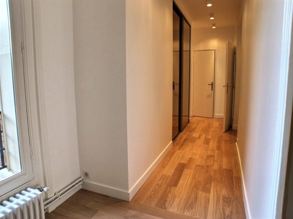 Cette image montre un couloir avec un mur blanc et un sol en bois brun.
