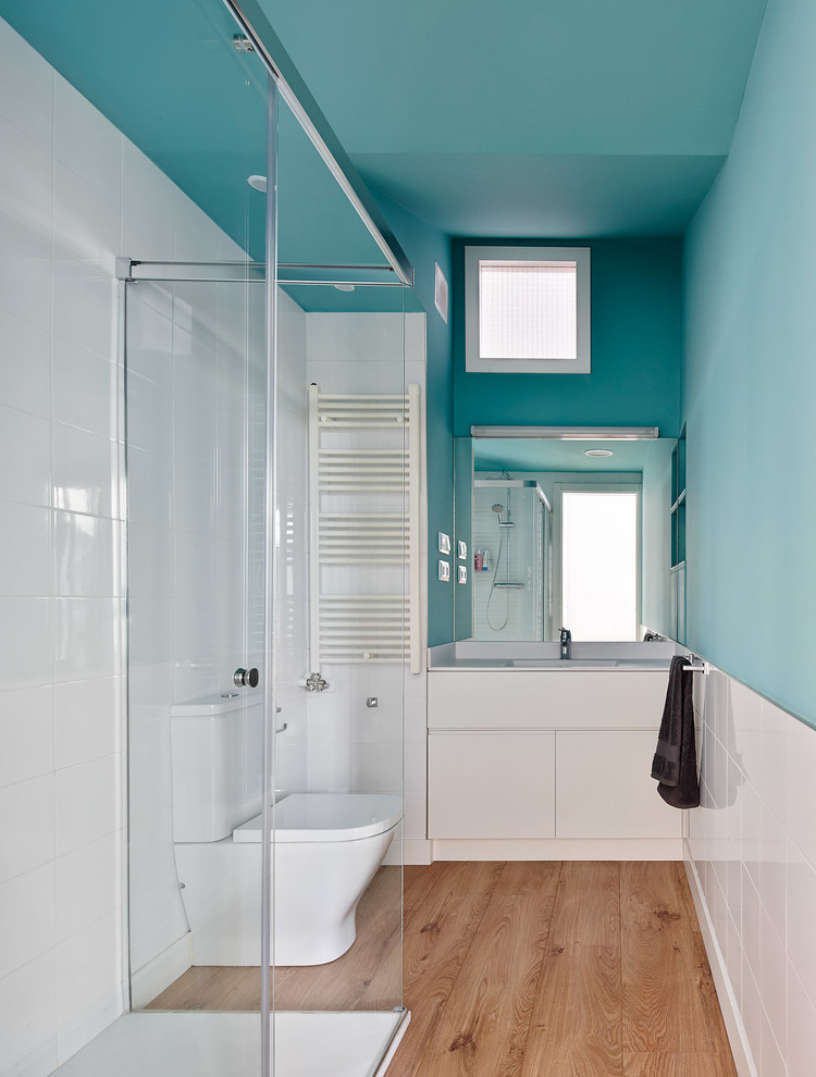 Diseño de cuarto de baño costero pequeño con armarios con paneles lisos, puertas de armario blancas, ducha esquinera, sanitario de dos piezas, paredes azules, suelo de madera en tonos medios, aseo y ducha y lavabo integrado