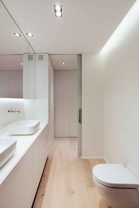 Réalisation d'une salle de bain minimaliste de taille moyenne avec un placard à porte plane, WC suspendus, un mur blanc, parquet clair, une vasque, un plan de toilette en surface solide et un plan de toilette blanc.