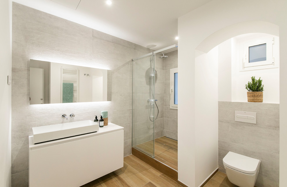 Mediterranean bathroom in Other with flat-panel cabinets, white cabinets, a wall mounted toilet, grey tiles, white walls, a vessel sink, brown floors, white worktops and a sliding door.