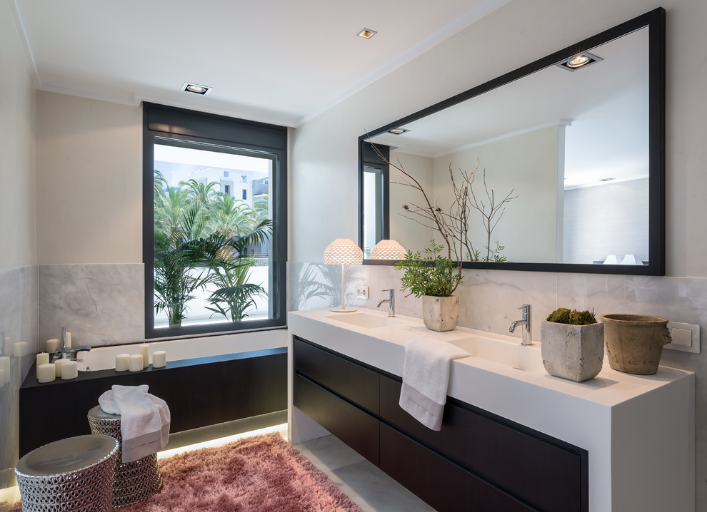 Immagine di una stanza da bagno design con ante lisce, ante nere, vasca sottopiano, piastrelle grigie, pareti bianche, lavabo integrato, pavimento grigio e top bianco