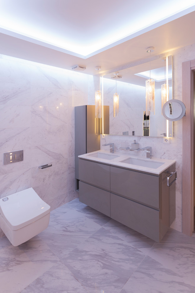 Пример оригинального дизайна: ванная комната в современном стиле с серыми фасадами, инсталляцией, белой плиткой, бежевой плиткой, серой плиткой, плоскими фасадами, серыми стенами, серым полом, белой столешницей, врезной раковиной и зеркалом с подсветкой