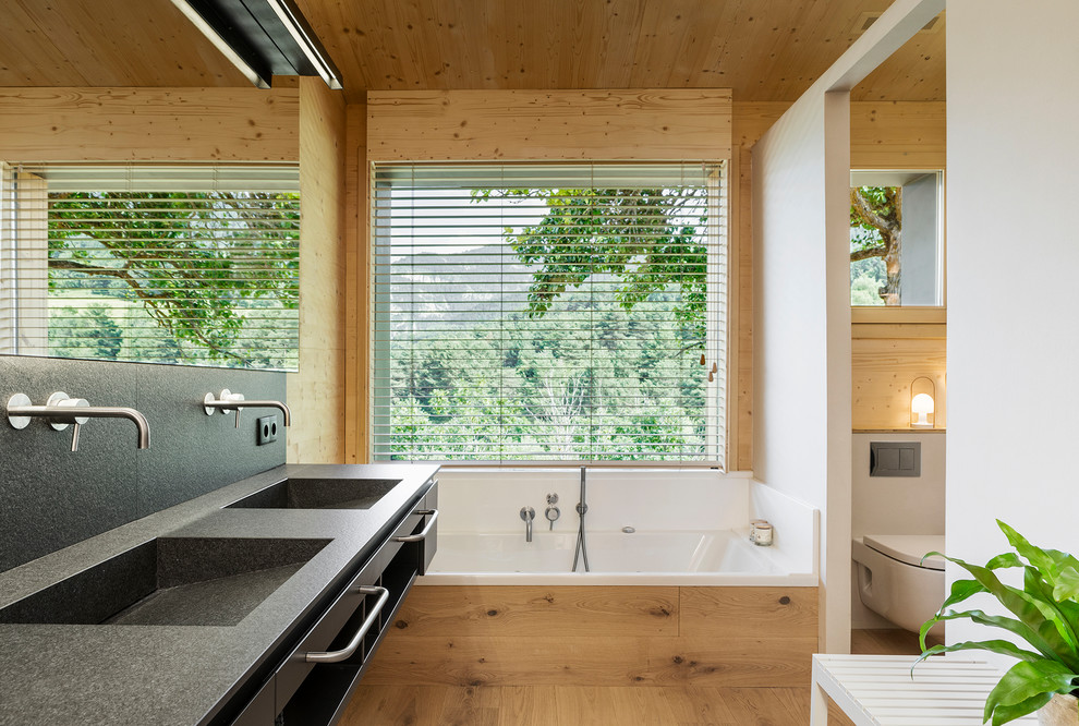 Modern inredning av ett svart svart en-suite badrum, med släta luckor, svarta skåp, ett badkar i en alkov, en vägghängd toalettstol, vita väggar, ljust trägolv och ett integrerad handfat