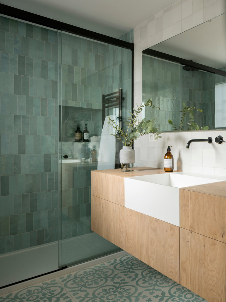 Modelo de cuarto de baño escandinavo, de tamaño medio, con armarios con paneles lisos, puertas de armario de madera clara, ducha empotrada, baldosas y/o azulejos verdes, aseo y ducha, encimera de madera, suelo multicolor y ducha con puerta corredera