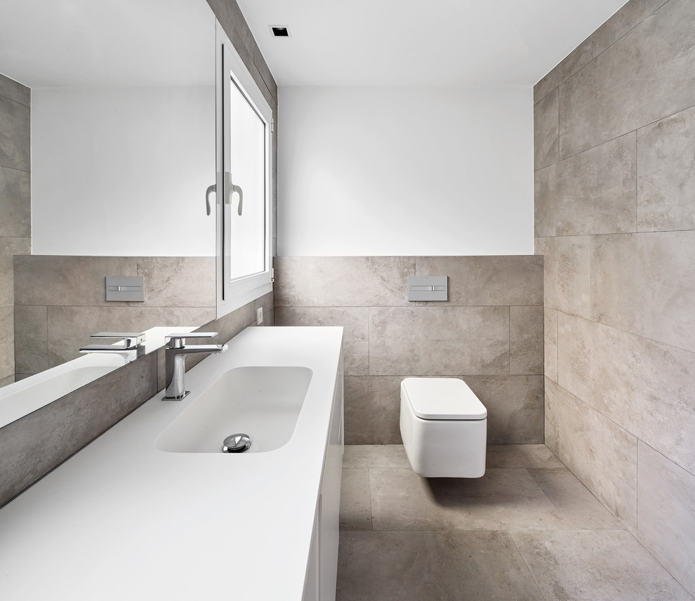 Modernes Badezimmer mit weißen Schränken, integriertem Waschbecken, flächenbündigen Schrankfronten, Wandtoilette, weißer Wandfarbe, weißer Waschtischplatte, grauen Fliesen und grauem Boden in Sonstige