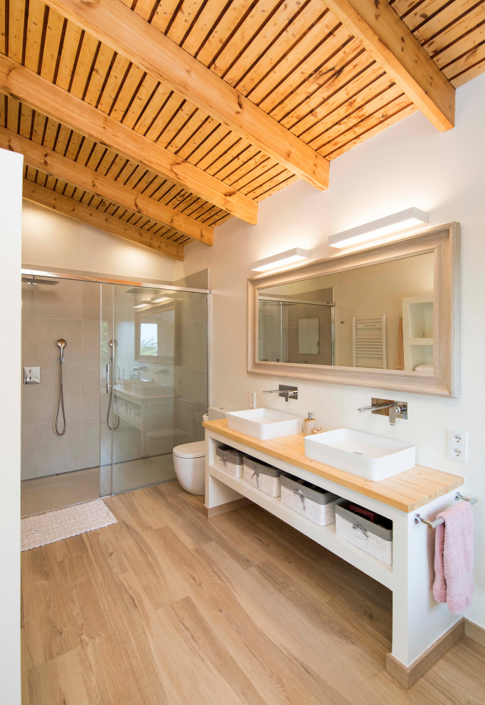 Foto på ett medelhavsstil beige badrum, med öppna hyllor, vita skåp, en kantlös dusch, grå kakel, vita väggar, ljust trägolv, ett fristående handfat, träbänkskiva, beiget golv och dusch med skjutdörr