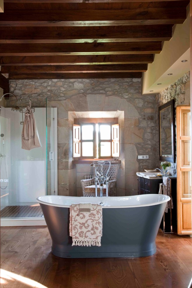 Modelo de cuarto de baño mediterráneo con bañera exenta, ducha esquinera, suelo de madera en tonos medios, suelo marrón, ducha con puerta con bisagras y piedra