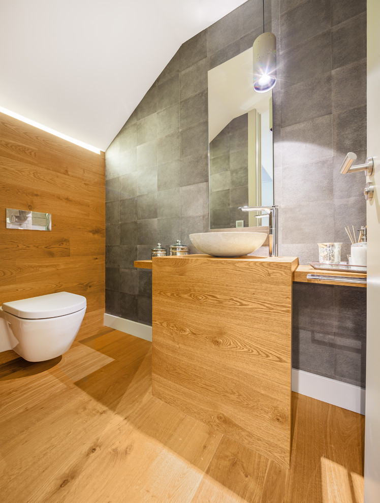 Modern inredning av ett mellanstort badrum med dusch, med ett fristående handfat, träbänkskiva, grå kakel, mellanmörkt trägolv, en vägghängd toalettstol och grå väggar