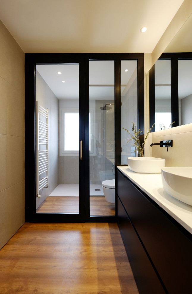 Cette image montre une grande salle d'eau design avec un placard à porte plane, des portes de placard noires, une douche ouverte, WC suspendus, un carrelage blanc, des carreaux de porcelaine, un sol en bois brun, une vasque, un sol marron et un plan de toilette blanc.