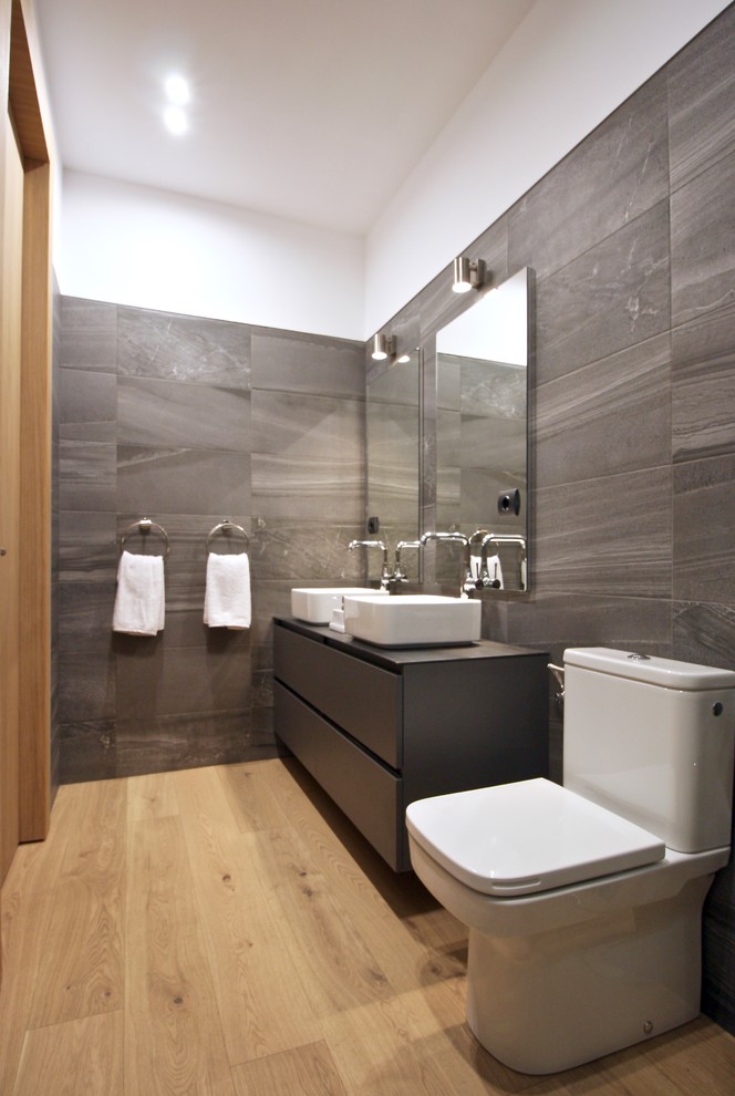 Imagen de cuarto de baño contemporáneo con puertas de armario grises, baldosas y/o azulejos grises, paredes grises, suelo de madera en tonos medios, lavabo sobreencimera y armarios con paneles lisos