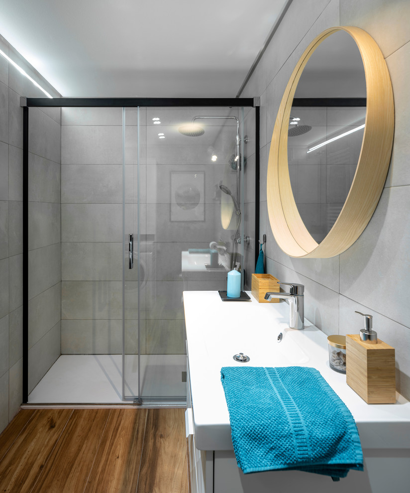 Inspiration for a medium sized scandi ensuite bathroom in Bilbao with raised-panel cabinets, white cabinets, a built-in shower, a one-piece toilet, grey tiles, ceramic tiles, grey walls, ceramic flooring, a trough sink, solid surface worktops, brown floors, a sliding door and white worktops.