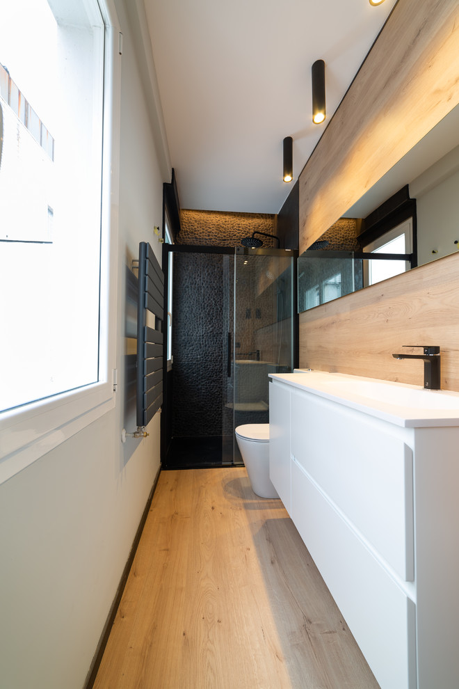 Стильный дизайн: ванная комната в современном стиле с плоскими фасадами, белыми фасадами, душем в нише, унитазом-моноблоком, черной плиткой, галечной плиткой, серыми стенами, светлым паркетным полом, душевой кабиной, монолитной раковиной, бежевым полом, душем с раздвижными дверями и белой столешницей - последний тренд