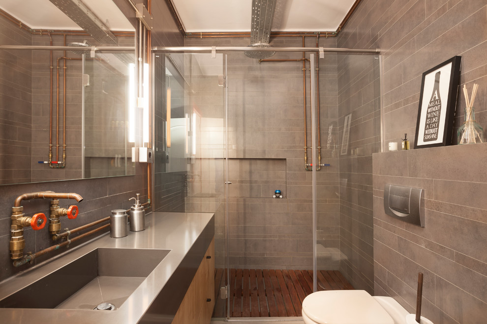 Industriell inredning av ett grå grått badrum med dusch, med släta luckor, skåp i ljust trä, en dusch i en alkov, en toalettstol med hel cisternkåpa, grå kakel, ett integrerad handfat, bänkskiva i rostfritt stål och dusch med skjutdörr