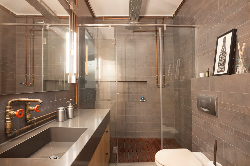 Exemple d'une salle de bain industrielle en bois brun de taille moyenne avec un placard à porte plane, WC à poser, un carrelage gris, des carreaux de céramique, un lavabo intégré, un plan de toilette en acier inoxydable et une cabine de douche à porte battante.