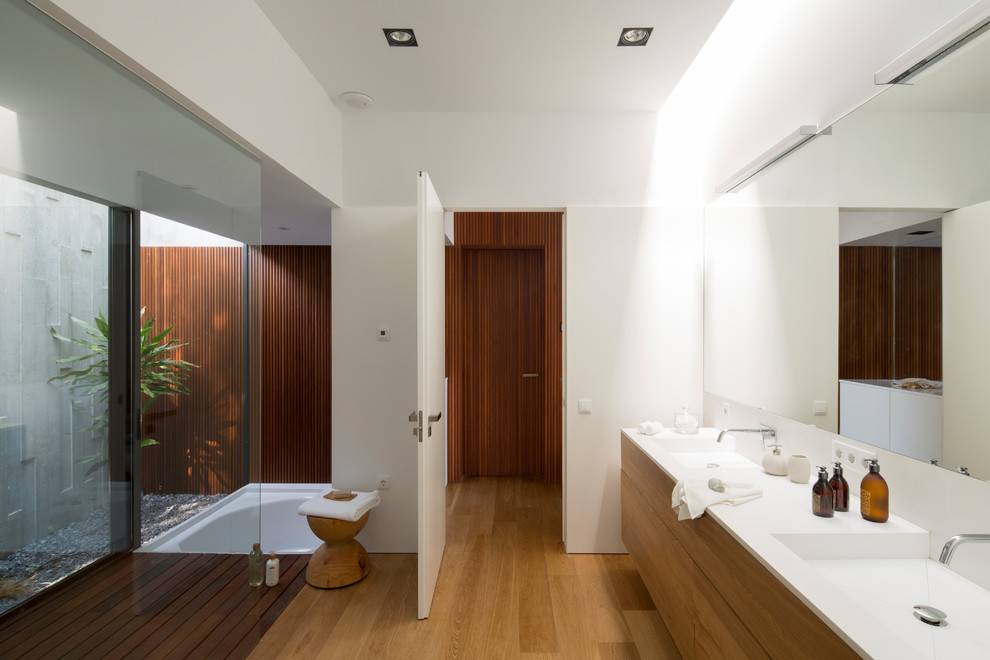 Ejemplo de cuarto de baño actual con suelo de madera en tonos medios, suelo marrón, armarios con paneles lisos, puertas de armario de madera oscura, paredes blancas, lavabo integrado y encimeras blancas
