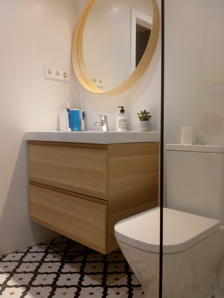 Nordisk inredning av ett mellanstort vit vitt badrum med dusch, med släta luckor, vita skåp, en kantlös dusch, en vägghängd toalettstol, keramikplattor, vita väggar, mosaikgolv, ett väggmonterat handfat och flerfärgat golv