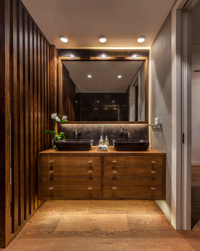 Cette photo montre une salle d'eau tendance en bois brun de taille moyenne avec un placard à porte plane, un carrelage noir, un carrelage gris, un mur gris, un sol en bois brun, une vasque, un plan de toilette en bois, un sol marron et un plan de toilette marron.