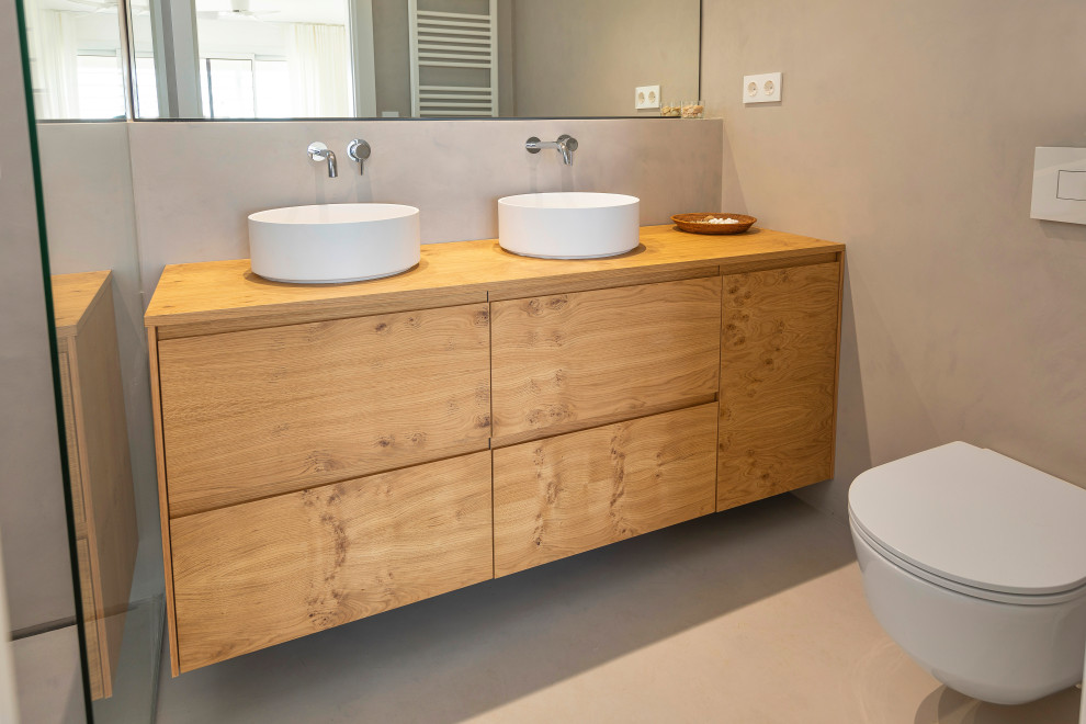This is an example of a large mediterranean ensuite bathroom in Other with freestanding cabinets, medium wood cabinets, a built-in shower, a wall mounted toilet, grey tiles, grey walls, concrete flooring, a vessel sink, wooden worktops, grey floors and an open shower.