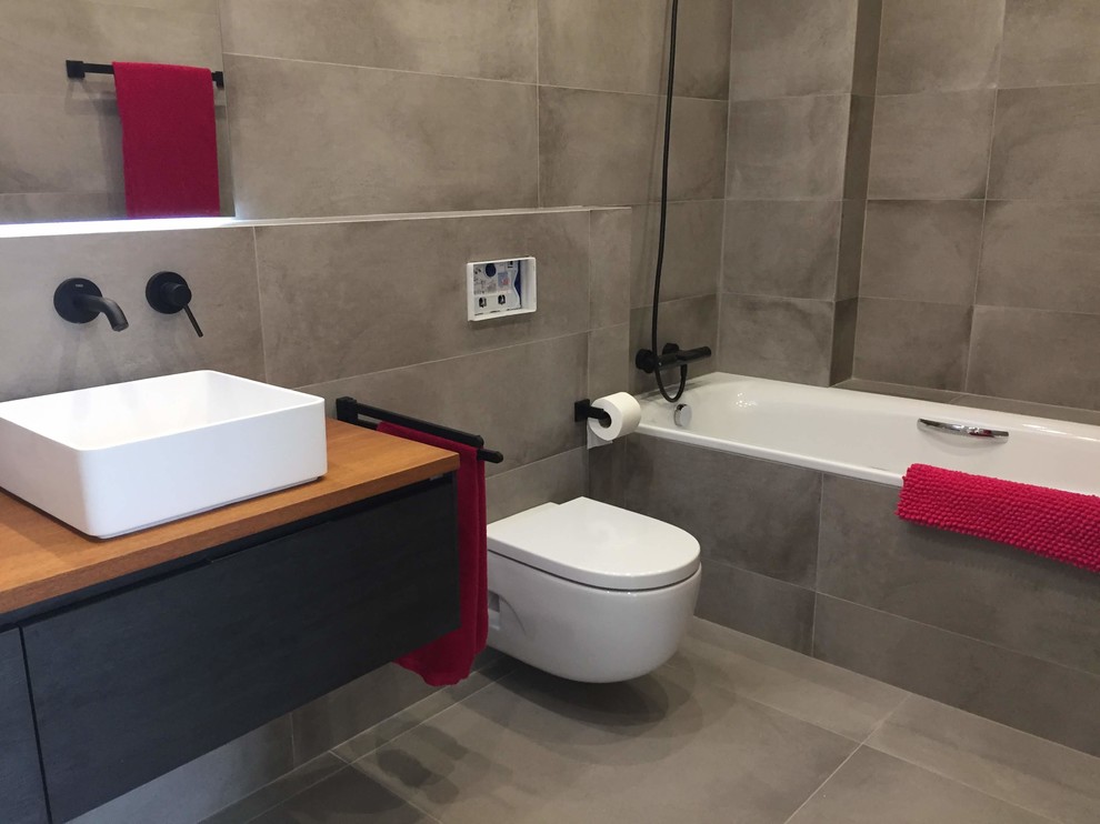 Photo of an urban ensuite bathroom in Barcelona with freestanding cabinets, black cabinets, an alcove bath, a wall mounted toilet, grey tiles, porcelain tiles, grey walls, ceramic flooring, a vessel sink, wooden worktops, grey floors and brown worktops.