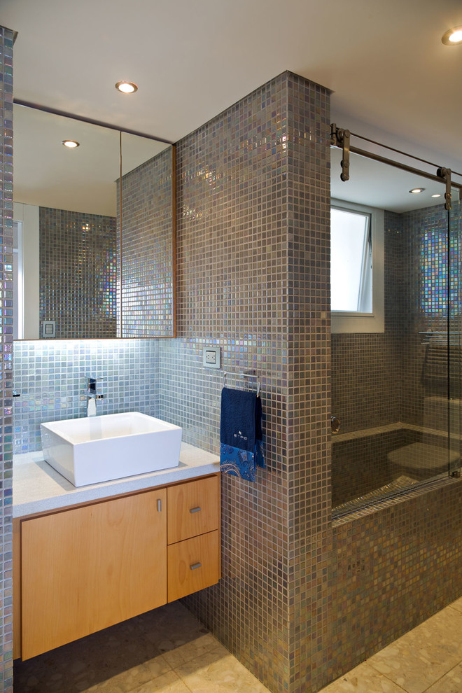 Aménagement d'une salle de bain contemporaine en bois brun avec un carrelage gris, des carreaux de miroir, une vasque, une cabine de douche à porte coulissante, un plan de toilette blanc, un placard à porte plane et un sol beige.