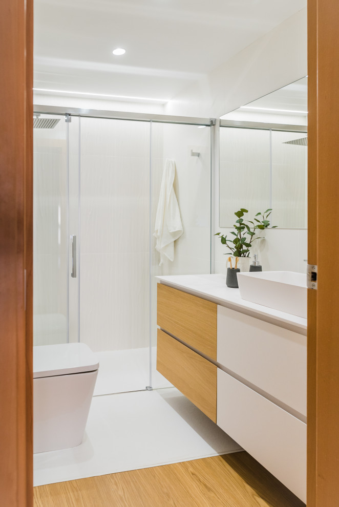 Cette image montre une salle de bain design de taille moyenne avec un placard à porte plane, des portes de placard blanches, WC à poser, un carrelage blanc, un mur blanc, un sol en bois brun, une vasque, un sol marron, une cabine de douche à porte coulissante et un plan de toilette blanc.