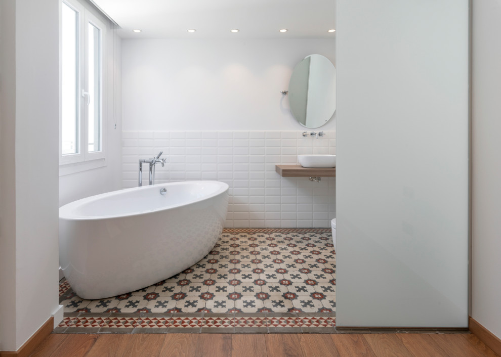 Bild på ett mellanstort funkis beige beige en-suite badrum, med ett fristående badkar, vit kakel, porslinskakel, vita väggar, ett fristående handfat, träbänkskiva och flerfärgat golv