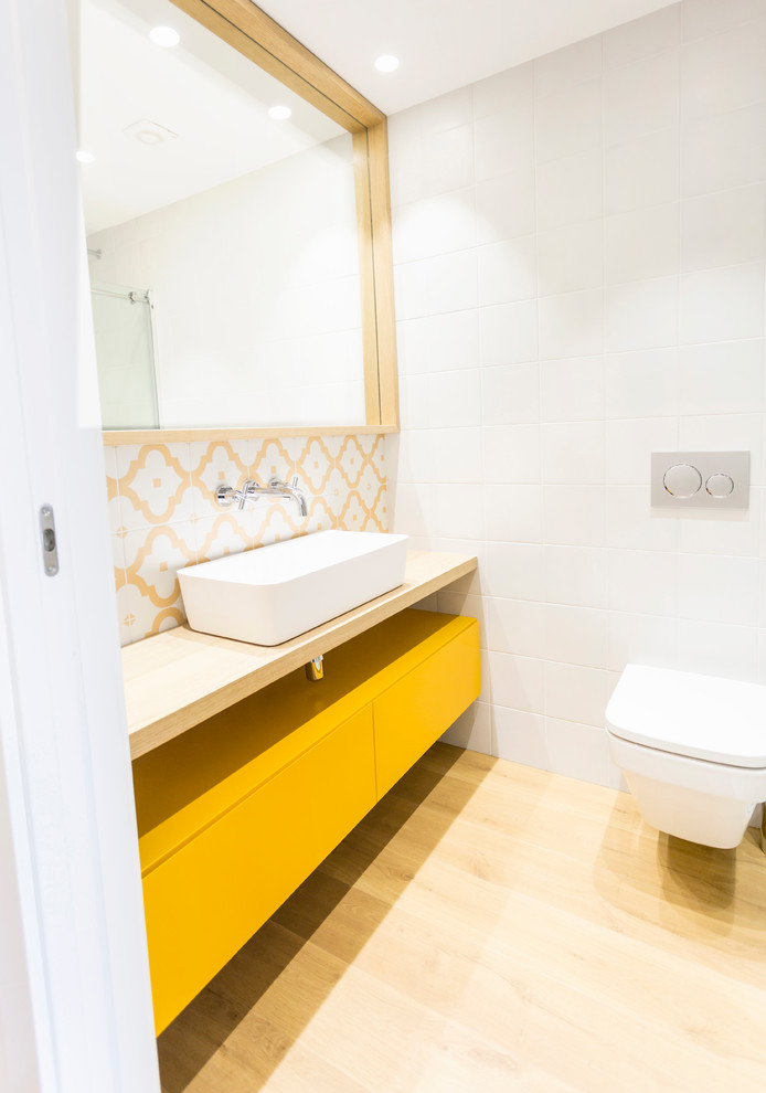 Cette photo montre une salle de bain scandinave avec des portes de placard jaunes, un mur blanc, un plan de toilette en bois, un placard à porte plane, WC suspendus, un carrelage multicolore, un carrelage blanc, un carrelage jaune, parquet clair et une vasque.