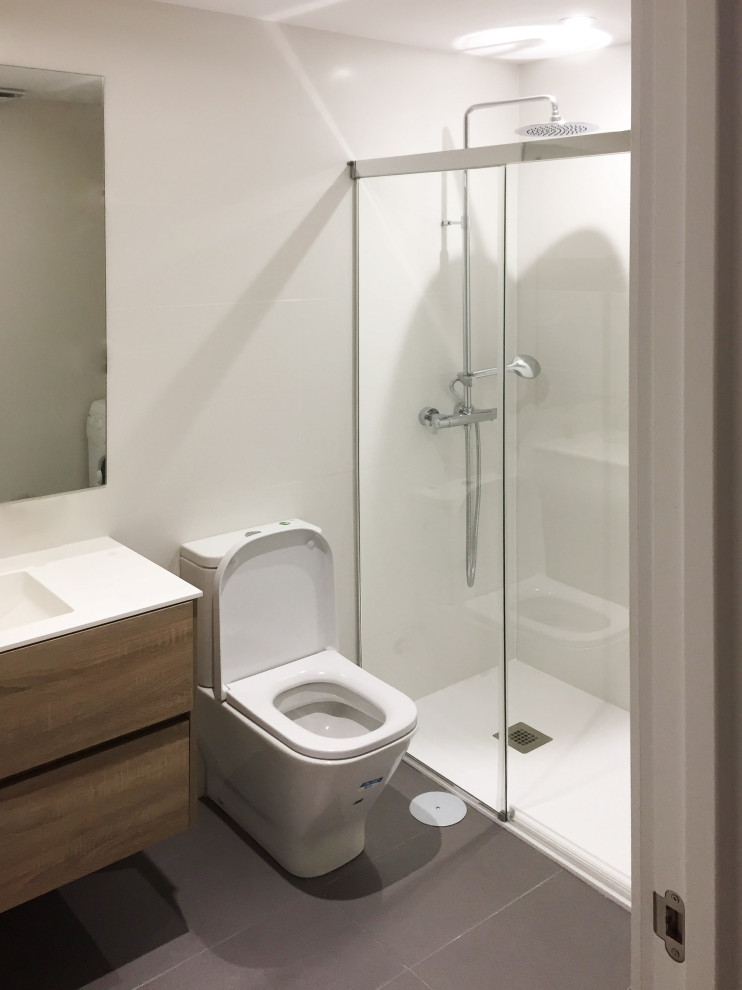 Modern inredning av ett litet en-suite badrum, med bruna skåp, en toalettstol med separat cisternkåpa, keramikplattor, vita väggar, klinkergolv i keramik, ett väggmonterat handfat, bänkskiva i akrylsten, grått golv, möbel-liknande, en kantlös dusch, vit kakel och dusch med skjutdörr