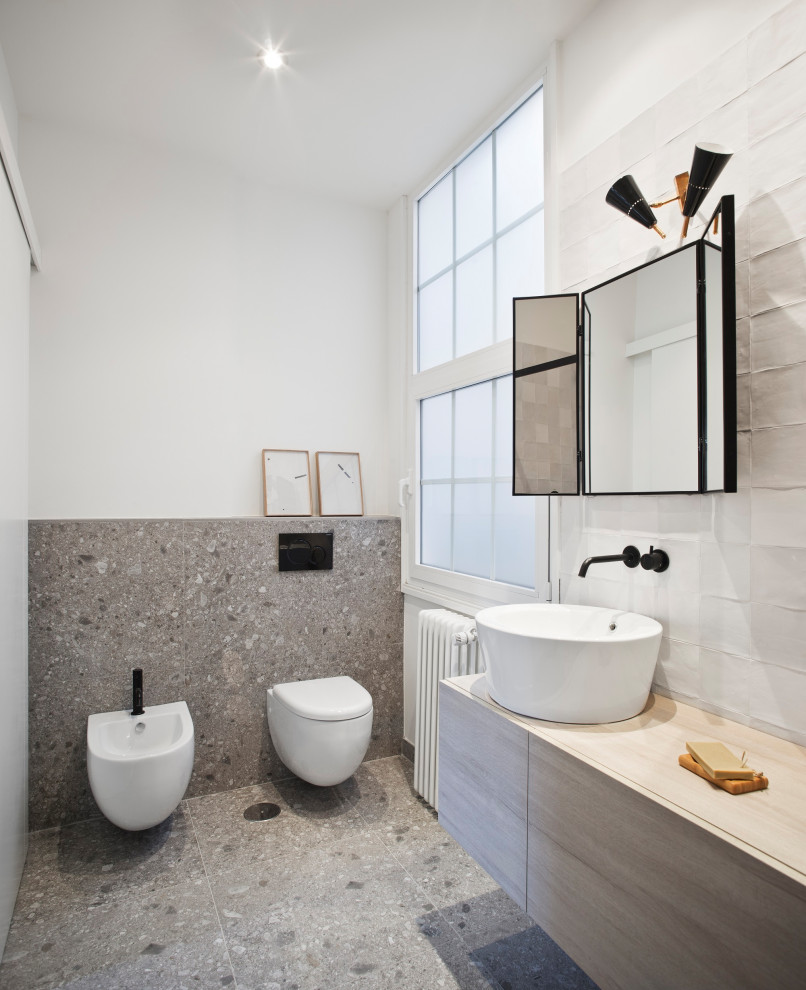 Cette photo montre une salle de bain tendance avec un placard à porte plane, des portes de placard grises, un bidet, un carrelage blanc, un mur blanc, un sol en terrazzo, une vasque et un sol gris.