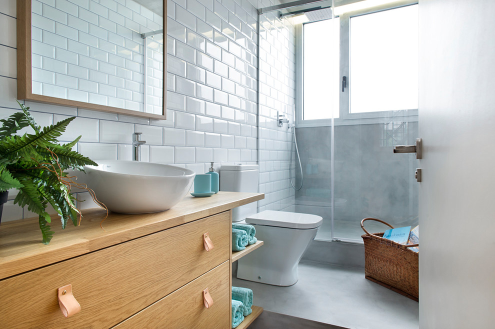 Foto på ett industriellt badrum med dusch, med släta luckor, skåp i ljust trä, en dusch i en alkov, en toalettstol med separat cisternkåpa, vit kakel, vita väggar, betonggolv, ett fristående handfat, träbänkskiva och grått golv