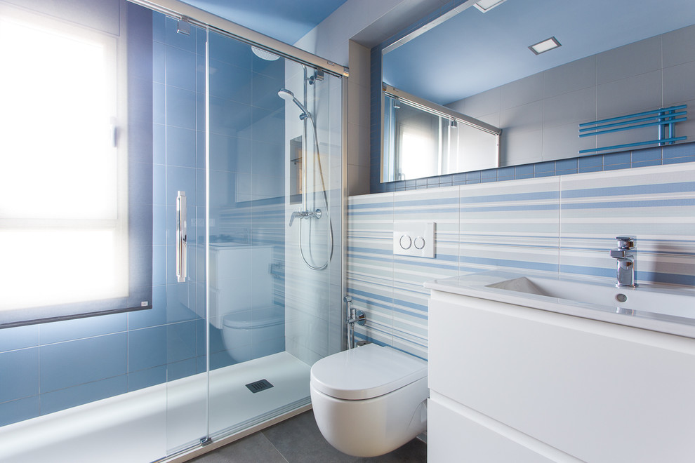 Diseño de cuarto de baño costero de tamaño medio con armarios con paneles lisos, puertas de armario blancas, ducha empotrada, sanitario de pared, paredes azules, suelo de baldosas de cerámica, aseo y ducha y lavabo integrado
