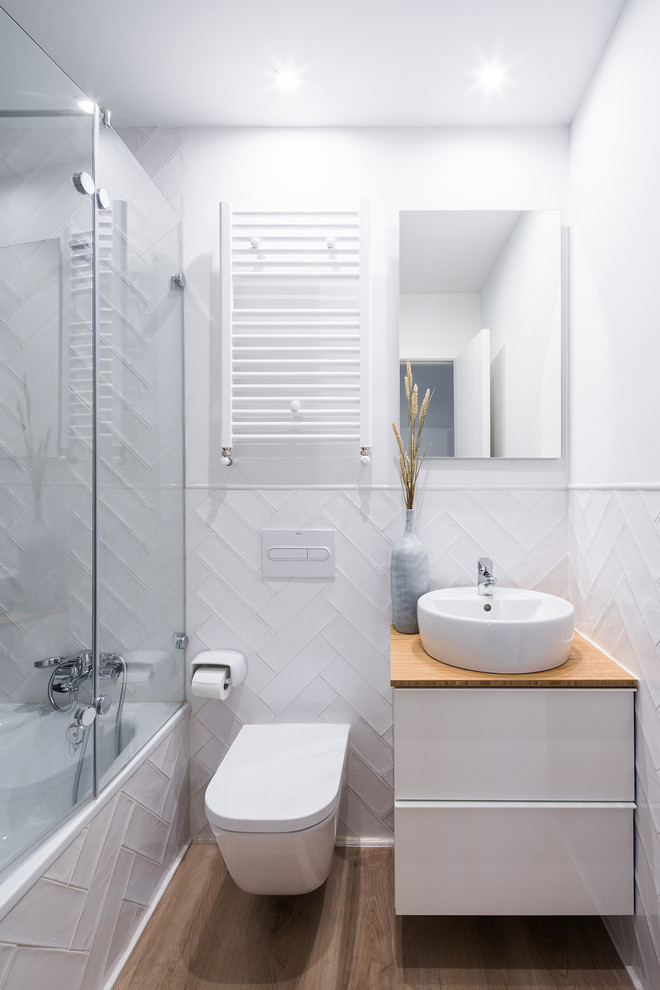 Nordisk inredning av ett brun brunt badrum med dusch, med släta luckor, vita skåp, ett badkar i en alkov, en dusch/badkar-kombination, en vägghängd toalettstol, vit kakel, vita väggar, ett fristående handfat, träbänkskiva, brunt golv och dusch med gångjärnsdörr